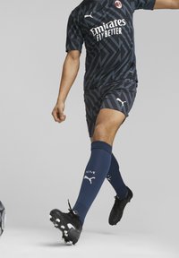 Tmavě modrý fotbalový dres s geometrickými vzory, který má logo na hrudi, v kombinaci s námořnickými ponožkami a černými kopačkami, zachycený v akci.