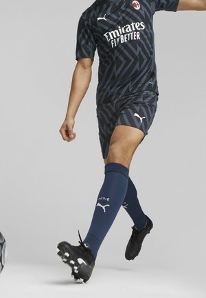 Équipement de football bleu foncé avec des motifs géométriques, arborant un logo sur la poitrine, associé à des chaussettes bleu marine et des crampons noirs, capturé en pleine action.