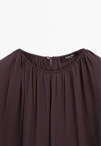 Blouse violet foncé avec encolure froncée et petite fermeture bouton au dos, avec une étiquette de marque visible à l'intérieur du col.