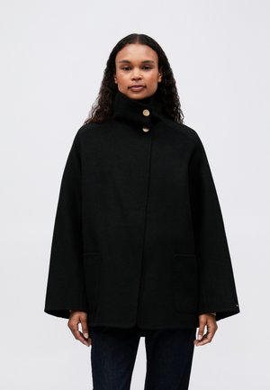 Vrouw in een zwart cape-jasje met hoge kraag, gouden knopen, voorzakken en lange wijde mouwen, staande tegen een effen achtergrond.