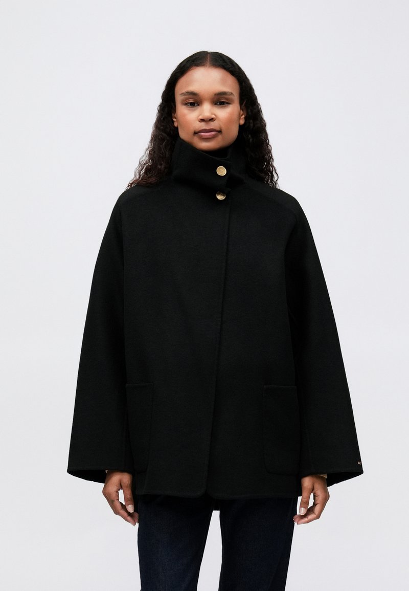 Kvinde iført en sort kappejakke med høj krave, guldknapper, frontlommer og lange brede ærmer, stående foran en ensfarvet baggrund.