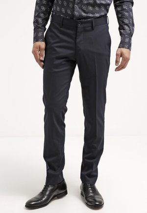 Homme portant un pantalon de costume noir ajusté, des chaussures en cuir noires et une chemise noire avec un motif géométrique blanc, debout devant un fond uni.