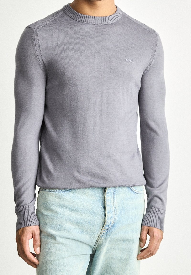 Homme portant un pull BOSS à manches longues gris clair et un jean bleu clair, debout devant un fond uni clair.