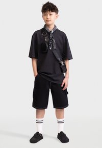 Schwarzes, übergroßes T-Shirt und passende Shorts, beide mit kontrastierenden Nähten. Accessoire-Tasche an einem Tragegurt, kombiniert mit gestreiften Socken und schwarzen Slippers.