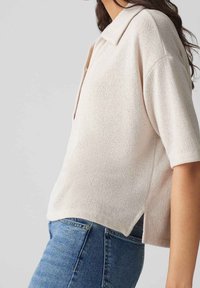 Kvinna iklädd en lös, kortärmad beige stickad polo t-shirt som kombineras med högmidjade blå jeans mot en enfärgad bakgrund.