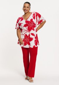 Witte top met rode bloemenpatronen en korte mouwen, gedragen met rode broek. De outfit heeft een losse fit en zachte stof.