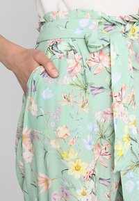 Kalhoty s květinovým vzorem v světle zelené barvě, vybavené vázáním v pase a postranními kapsami. Měkká, lehká látka s hladkým povrchem.