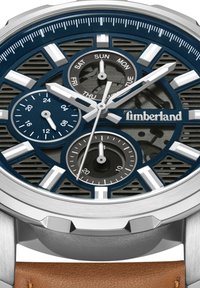 Timberland pulksteņi ar tumši zilu ciparnīcu, sudraba stundu apzīmējumiem un trim subciparnīcām. Iespējas ietver teksturētu korpusu un brūnu ādas siksnu.