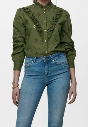 Vrouw draagt een olijfgroene blouse met ruches en gaatjesdetails en hoge blauwe spijkerbroek, staand tegen een effen achtergrond.