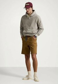 Mladý muž s brýlemi, červenou čepicí, šedou mikinou s kapucí, hnědými šortkami s opaskem, béžovými ponožkami a bílými teniskami, stojící s rukama v kapsách.