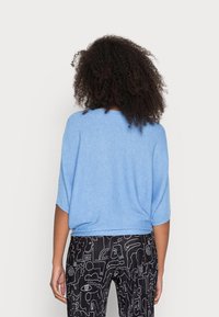 Hellblauer locker sitzender Pullover mit kurzen Dolman-Ärmeln, strukturiertem Stoff, kombiniert mit schwarzen gemusterten Hosen mit abstrakten weißen Mustern.