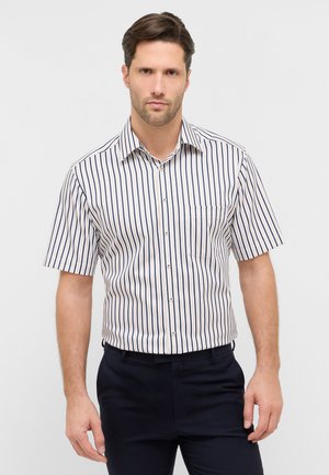 Mand iført en kortærmet, hvid skjorte med lodrette marineblå striber og mørke bukser, stående mod en ensfarvet lys baggrund.