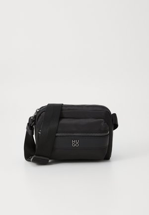 Schwarze rechteckige Crossbody-Tasche mit verstellbarem Riemen, Frontreißverschlusstasche und "HUGO" Logo auf dem Frontpanel vor weißem Hintergrund.