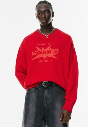 Red knitted sweater featuring the text "WANT IT DREAM IT GO FOR IT" in a contrasting design. Paired with high-waisted dark denim jeans.
