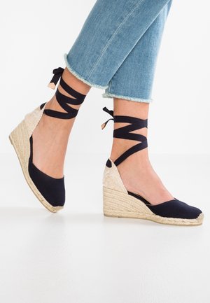 Wedge-Espadrilles mit einem Obermaterial aus marineblauem Stoff, beigem geflochtenem Absatz und schwarzen Knöchelbandagen. Das Design zeichnet sich durch eine abgerundete Spitze und eine subtile Textur aus.