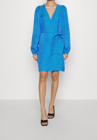 Mujer con un vestido envolvente texturizado de un brillante azul, con mangas largas abullonadas y sandalias de tacón negro y punta abierta, de pie frente a un fondo blanco.