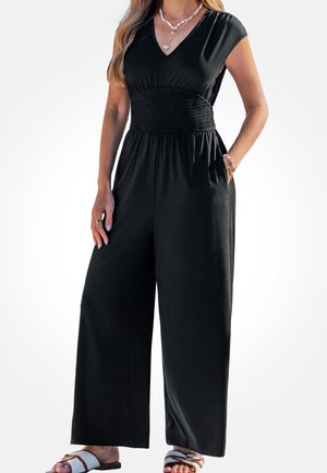 Femme portant une combinaison noire sans manches avec jambes larges, col en V, taille smockée, colliers superposés, bracelets et sandales blanches.