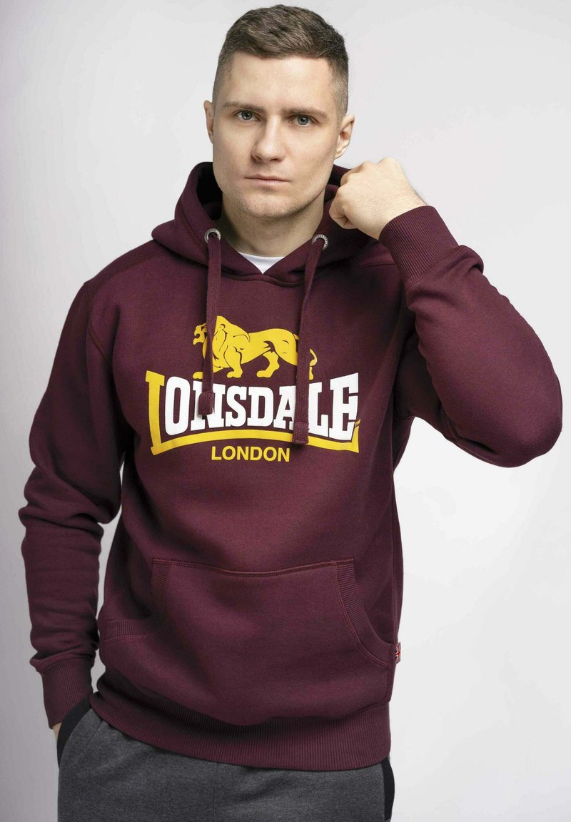 Felpa color bordeaux con logo di un leone giallo e testo bianco che recita "Lonsdale London." Presenta una tasca a marsupio frontale e cappuccio con cordino.