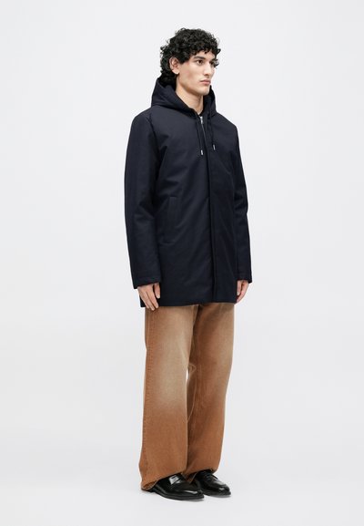 Meresinine kapuutsiga jakk, millel on esiosal tõmblukk, kaks küljetaskut ja mugav lõige, kombineeritud beežide laiade pükstega ja mustade jalanõudega.