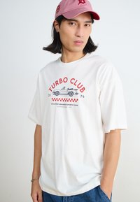 Balta medvilninė marškinėliai su „TURBO CLUB“ grafika, kurioje pavaizduotas senovinis kabrioletas, derinami su mėlynu džinsų kelnių ir rožine kepure.