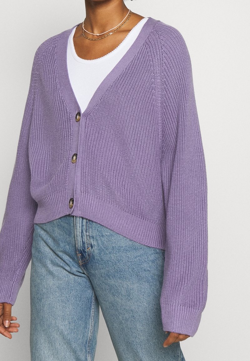 Lila stickad cardigan med V-ringning, knäppning och ribbade manschetter, tillsammans med ljusblå jeans. Texturerad tyg och kortad design.