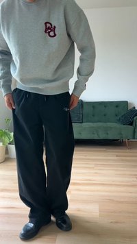 Γκρίζα φούτερ με κόκκινο λογότυπο, φαρδιά μαύρα παντελόνια και μαύρα παπούτσια. Το φόντο διαθέτει έναν πράσινο καναπέ και ένα φυτό.