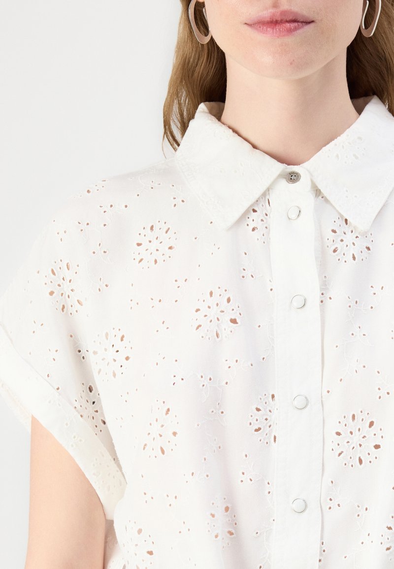 Camisa blanca de botones con mangas cortas, que presenta bordados de ojal florales, un cuello con solapa y cierres de botones plateados.
