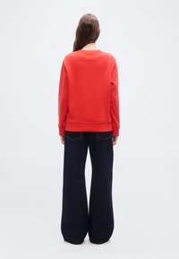 Άτομο με μακριά μαλλιά στέκεται με την πλάτη γυρισμένη, φορώντας κόκκινο φούτερ και μπλε σκουρόχρωμο τζιν με φαρδιά μπατζάκια, μπροστά σε απλό φόντο.