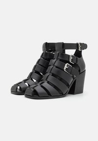 Sorte lærreimode sandaler med åpen tå, med brede stropper og justerbare ankelspenner. Strukturert overflate og blokkhelldesign.