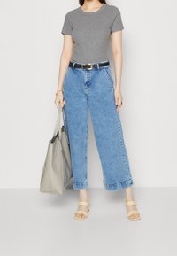 Nainen, jolla on harmaa t-paita, korkeavyötäröiset leveät siniset farkut, beiget korkokengät ja suuri harmaa olkalaukku.