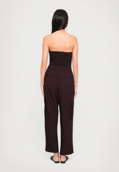 Pánt nélküli bordó overál, smocked szívvel, laza szabással, széles lábakkal és oldalsó cargo zsebekkel. Fekete slip-on szandálok láthatók.