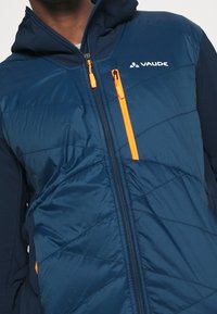 Marinblå isolerad jacka med huva, med orange dragkedje detaljer och diagonalt kviltad textur. Logotypen synlig på bröstet.