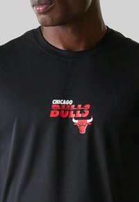 Mann trägt ein schwarzes T-Shirt mit rotem und weißem Chicago Bulls-Logo und Bullenkopf-Emblem auf der Brust.