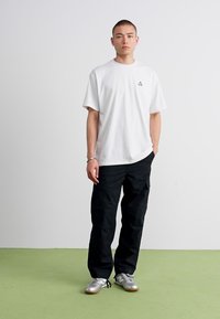 Young man standing against white wall wearing white oversized t-shirt, black cargo pants, and silver sneakers on a light green floor.