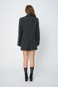 Blazer grigio a righe con vestibilità su misura, dotato di una fessura sul retro e bottoni metallici sui polsini. Indossato sopra una mini gonna plissettata.