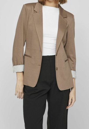 Mujer con el pelo castaño hasta la barbilla lleva una blazer beige, camisa blanca y pantalones negros. Las mangas de la blazer están remangadas, mostrando el forro a rayas.