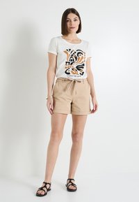 Pantalones cortos beige con dobladillos enrollados, combinados con una camiseta crema que presenta un diseño abstracto en negro y naranja. La modelo está de pie sobre un fondo blanco.