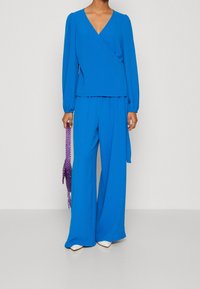 Blue wrap top with long sleeves paired with wide-leg pants. Smooth, textured fabric, held with a purple beaded accessory.