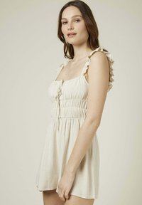 Robe beige clair en tissu texturé, présentant un corsage ajusté, des bretelles volantées et une taille élastique, se terminant au-dessus de la mi-cuisse.