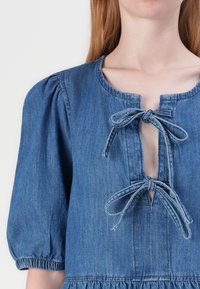 Denimkjole med puffermer, rund hals og en frontutsletting med knytninger. Stoffet har en jevn tekstur og en lys blå nyanse.