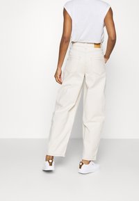 Krämmfärgade högmidjade byxor med avslappnad passform, utrustade med bakfickor och en vävd etikett vid midjan, kombinerade med vita sneakers.