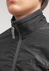 Un jeune portant une veste zippée noire avec un col relevé et un logo "G RAW" sur la poitrine.