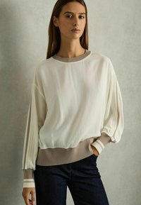 Ljus elfenbensfärgad pullover med långa, draperade ärmar och en ribbad beige nederkant. Har subtila ränder på ärmarna och en texturerad yta. Denimjeans synliga.