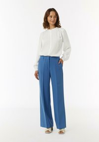 Biała bluzka z długim rękawem i okrągłym dekoltem, zestawiona z niebieskimi spodniami o szerokich nogawkach i wysokim stanem oraz jasnymi sandałami na obcasie.