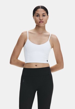 Donna con capelli scuri, canotta bianca corta e leggings neri a vita alta, in piedi con le mani dietro la schiena su sfondo semplice.