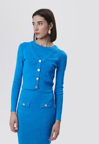 Cardigan blu lavorato a maglia con una forma boxy, scollo a V profondo, cinque bottoni dorati e una gonna abbinata con due tasche frontali e texture a motivo.