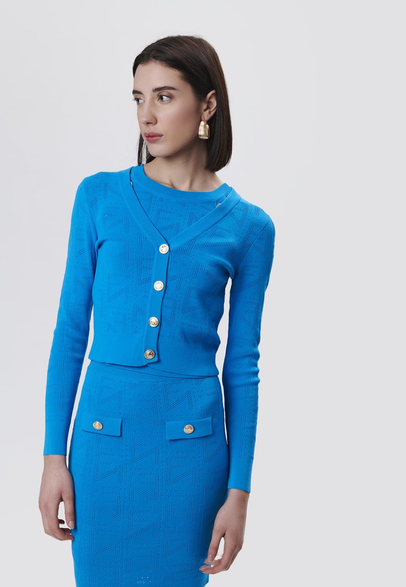 Cardigan blu lavorato a maglia con una forma boxy, scollo a V profondo, cinque bottoni dorati e una gonna abbinata con due tasche frontali e texture a motivo.