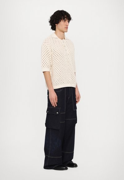 Polo en tricot, couleur blanc cassé, à manches courtes avec un tissage ouvert et un col côtelé, associé à un pantalon cargo en denim foncé avec plusieurs poches.