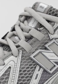 Szürke sportcipő hálós felsőrésszel, vastag fűzőkkel és fémes díszítésekkel. Figyelemre méltó részletei közé tartoznak a hatszögletű minták és a fényvisszaverő elemek.