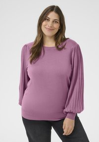 Leende kvinna med långt brunt hår, iklädd en mauve-tröja med veckade ballongsärmar och svarta byxor, som står mot en vit bakgrund.